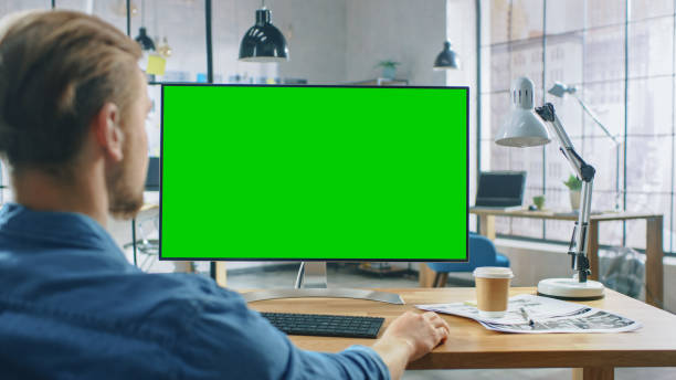 istock Over the Shoulder Shot: Man Uses Desktop Computer with Green Mock-up Screen while Sitting at His Desk. In the Background Stylish Modern Open Space Creative Business Office 1191658341