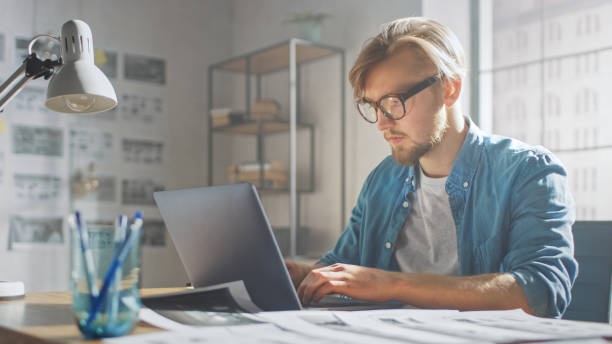 istock Creative Developer Works on a Laptop in His Studio. Designing Apps, Games and Doing Video Editing. In the Background Modern Sunny Loft Office with Project Storyboard and Concepts Covering the Wall 1191658368