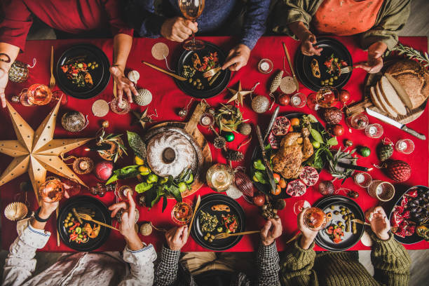Flat-lay of friends eating and talking at festive Christmas table Friends celebrating Christmas. Flat-lay of people eating and talking over festive table with red cloth with champagne, roasted chicken, bundt cake, fruits, decorations, top view. Winter holiday party winter food top view stock pictures, royalty-free photos & images