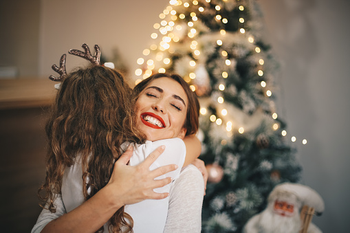 Beautiful mother smiling and hugging daughter wearing Christmas headband to celebrate New year's Eve. There's a Christmas tree behind.