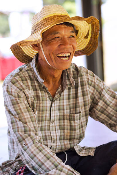 Sitting cheerfully laughing thai mature farmer man is sitting outdoors in small village Pharungmee in province Phitsanulok. Man is sitting with other village people who are relaxing and talking. Man is wearing a hat.