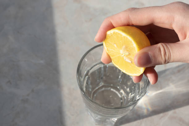 women hand squeezes lemon juice in glass with soda water on marble table background. detox healthy drink concept - lemon imagens e fotografias de stock