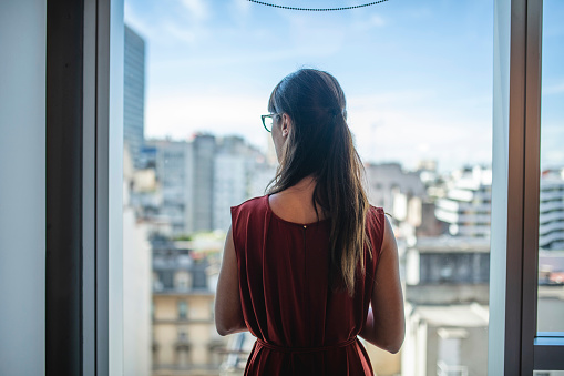 View from behind of longhaired businesswoman in mid 30s standing at window and looking out at downtown Buenos Aires.