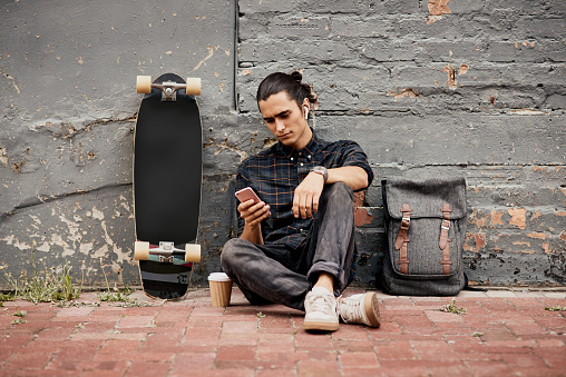 Full length shot of a handsome young man using his cellphone while sitting near his skateboard outside during the day