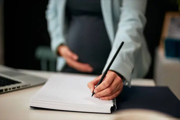 Caucasian pregnant woman in forties standing in office, writing down tasks and holding belly. Selective focus on hand.