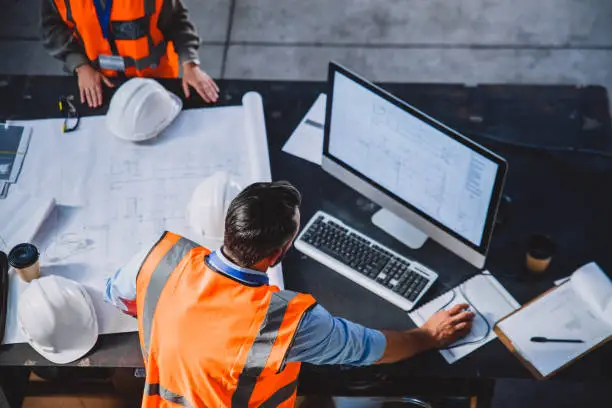 High angle shot of two engineers working together on blueprints while making use of a computer inside of a workshop