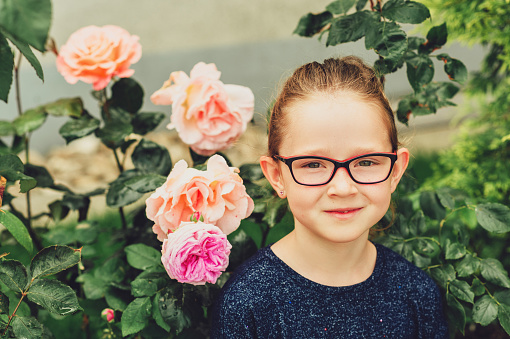 Summer portrait of pretty little girl wearing glasses, posing in rose garden