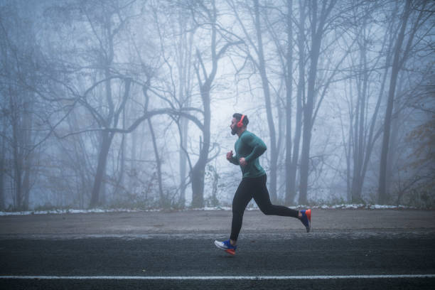 Man Running On The Road. Full length man wearing sports clothing and red headphones running on the road in foggy weather condition. sportsman professional sport side view horizontal stock pictures, royalty-free photos & images