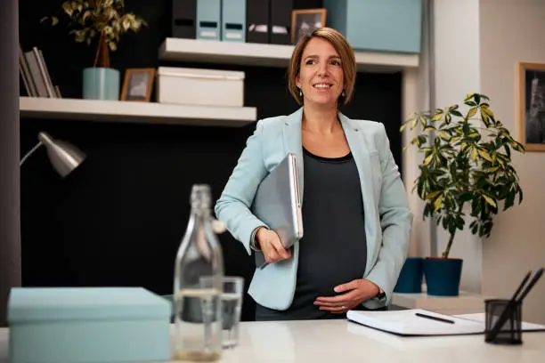 Positive smiling caucasian pregnant businesswoman holding laptop under armpit, touching belly and standing in office. She is preparing to go on business meeting with colleagues.