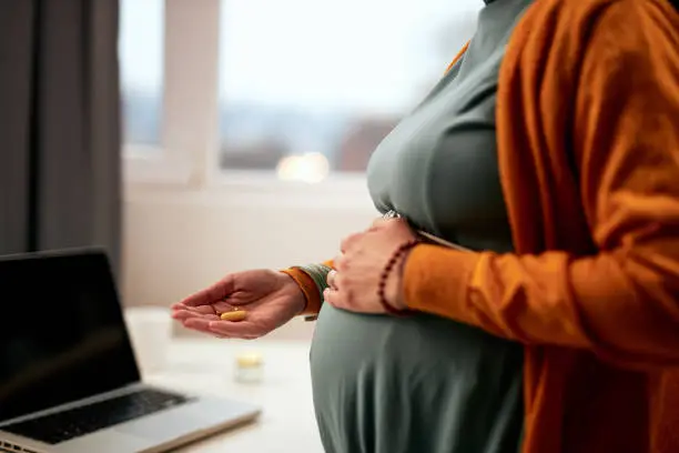 Cutout picture of pregnant caucasian businesswoman standing in office, touching belly and holding pill.