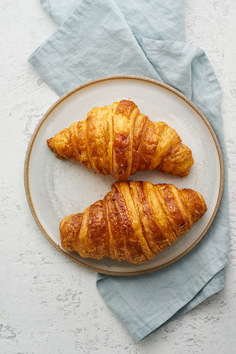 Two delicious croissants on a plate and a hot drink in a mug. Morning French breakfast with fresh pastries. Light gray background, vertical