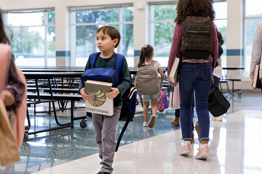 Hispanic elementary school student carries a math textbook as he walks in the school cafeteria.