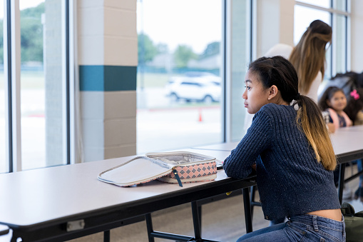 Lonely middle schoolgirl stares out the window while eating lunch in the school cafeteria.