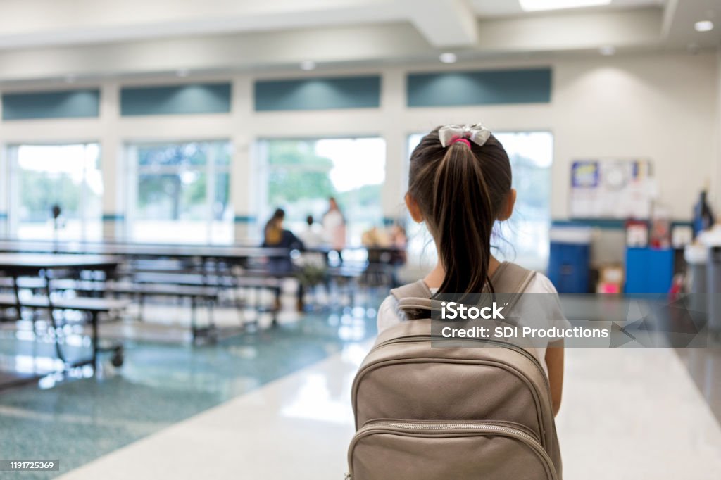 Rear view of young schoolgirl entering cafeteria Elementary schoolgirl enters the school cafeteria. She pauses while looking for a friend. School Building Stock Photo