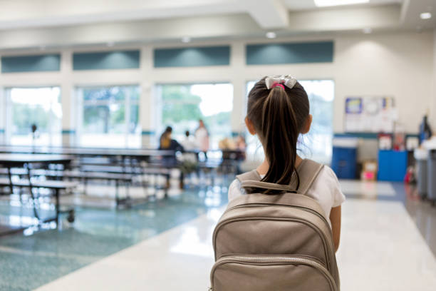 Rear view of young schoolgirl entering cafeteria Elementary schoolgirl enters the school cafeteria. She pauses while looking for a friend. bag photos stock pictures, royalty-free photos & images