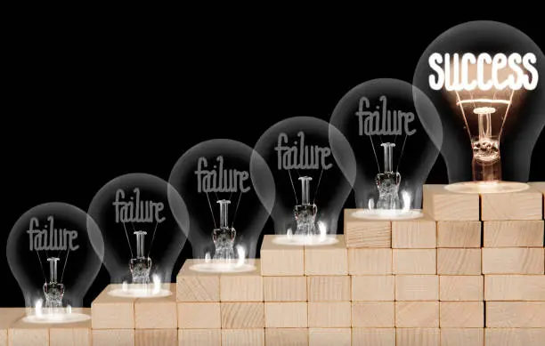 Group of shining and dimmed light bulbs on wooden block ladder with fibers in a shape of Failure and Success concept words isolated on black background.