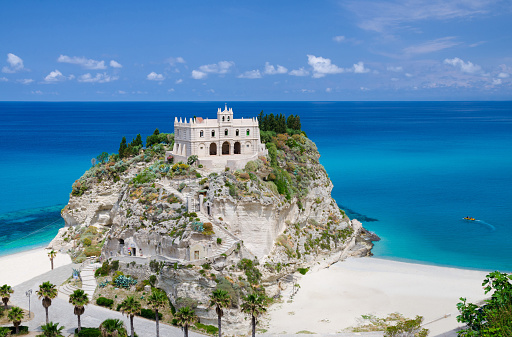 Monastery Sanctuary church of Santa Maria dell Isola on top of rock Tyrrhenian Sea and green palm trees, blue sky white clouds in summer clear day, Tropea town, Vibo Valentia, Calabria, Southern Italy