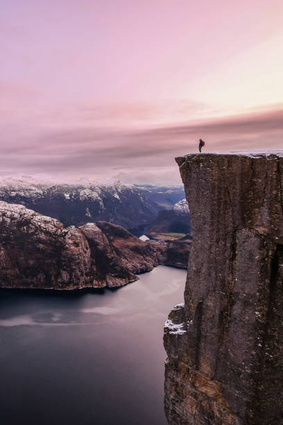 Man hiker standing on the famous Preikestolen over the Lysefjord, beautiful colors at sunset, Ryfylke, Rogaland, Norway Man hiker standing on the famous Preikestolen Pulpit Rock over the Lysefjord, some snow on the peaks and beautiful colors at sunset, Ryfylke, Rogaland, Norway, Scandinavia, Europe ryfylke stock pictures, royalty-free photos & images
