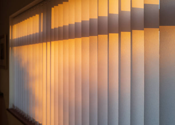 istock White textured vertical slat blinds hanging in front of a window as the sun is setting turning the light golden. 1191740474