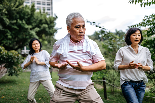 Group of three seniors taking an exercise class in the park, practicing tai chi, standing with their hands raised