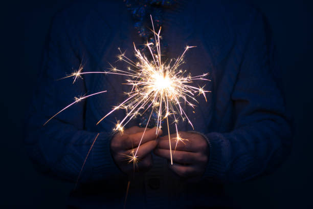 single man in grey knitted sweater and tinsel around his neck holding sparkler in dark at night