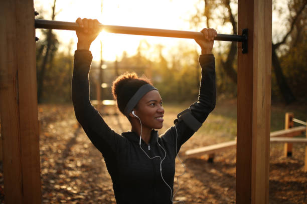 happy african american kobieta sportowiec robi podbródek na sprzęt do ćwiczeń o zachodzie słońca. - chin ups audio zdjęcia i obrazy z banku zdjęć