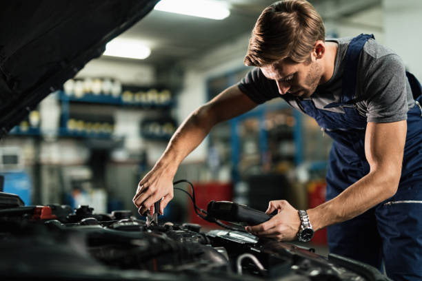 Auto mechanic using diagnostic tool while repairing car engine in a workshop. Young mechanic examining car battery with diagnostic tool while working in auto repair shop. jumper cable stock pictures, royalty-free photos & images