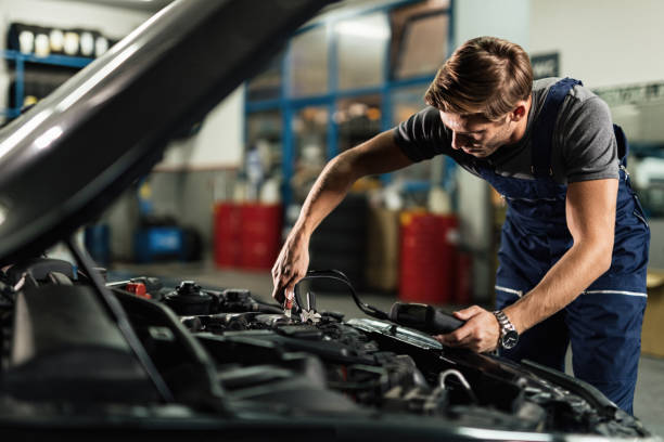 Young mechanic attaching jumper cables on car engine while working in auto repair shop. Auto mechanic using diagnostic tool and attaching jumper cables on car engine in a repair shop. car battery stock pictures, royalty-free photos & images