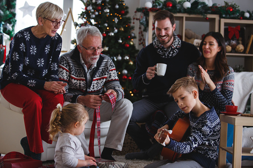 Three generation family enjoying Christmas day together. They are sitting by the Christmas tree in the living room. A boy is playing the guitar, his family is looking and listening to young performer.