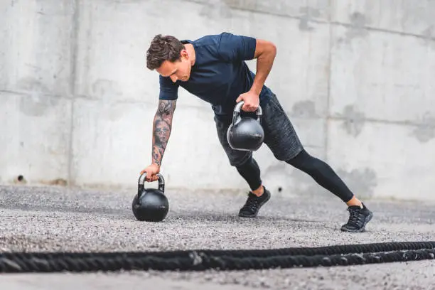 Low angle view of fit Caucasian male athlete in mid 20s doing kettlebell plank row exercise outdoors with battle ropes in foreground.