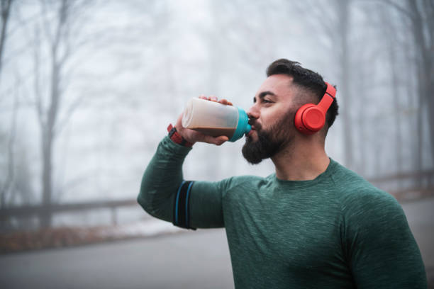 Man Drinking Energy Drink After Exercising. Sportsperson drinking protein after long exercising on the road, using wireless headphones. cocktail shaker photos stock pictures, royalty-free photos & images