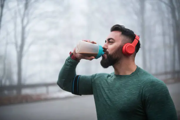 Sportsperson drinking protein after long exercising on the road, using wireless headphones.