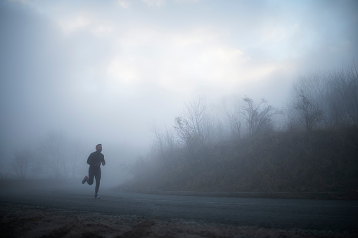 Full length man wearing sports clothing and red headphones running on the road in foggy weather condition.