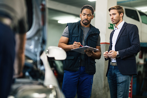 Young businessman and African American car mechanic talking while examining car at auto repair shop.