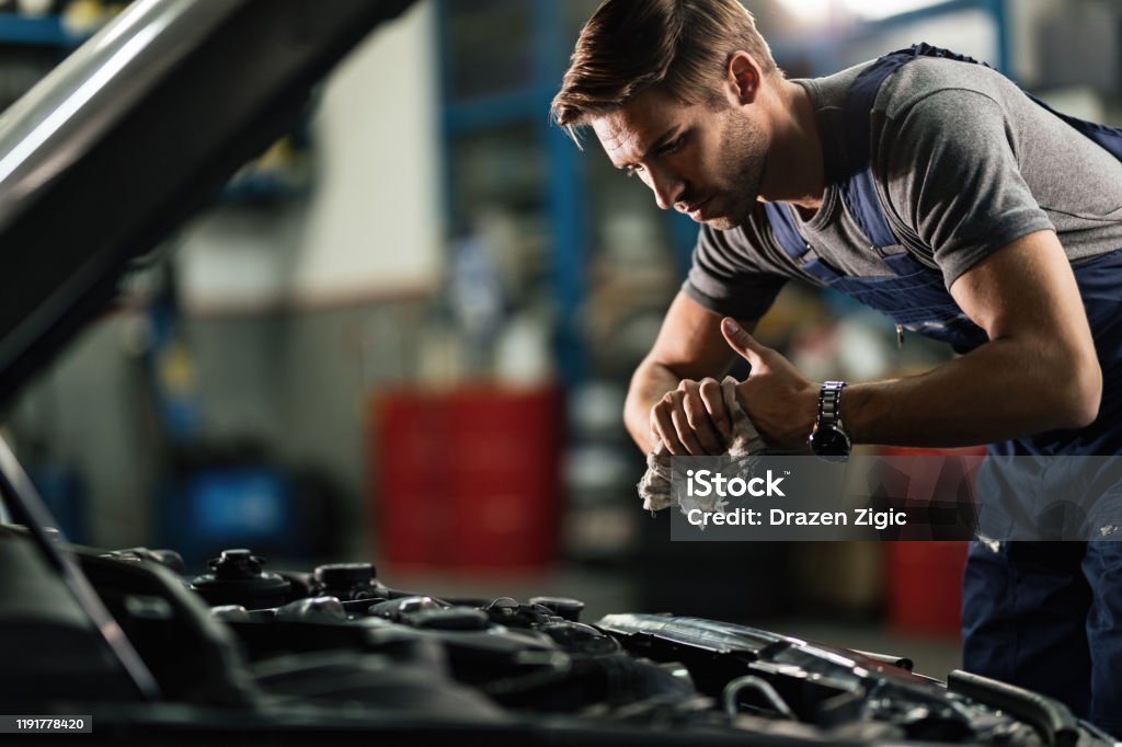 Young auto mechanic cleaning hands after working on car engine in a garage. Young mechanic wiping his hands while repairing car engine in auto repair shop. Auto Repair Shop Stock Photo