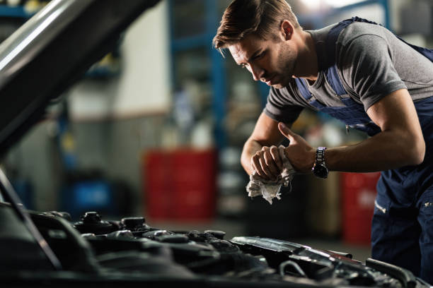 Young auto mechanic cleaning hands after working on car engine in a garage. Young mechanic wiping his hands while repairing car engine in auto repair shop. auto mechanic photos stock pictures, royalty-free photos & images