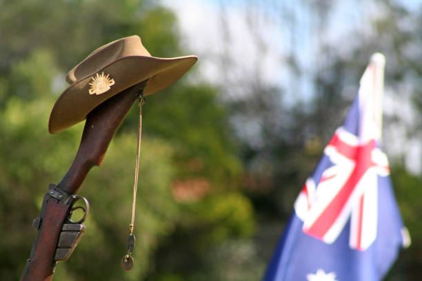 Close-up of an upturned Australian 303 rifle butt upon which sits a traditional Australian Slouch Hat with rising sun badge, dogtags and the Australian National Flag on ANZAC Day Landscape (with room for copy) of an upturned, vintage Australian Army 303 rifle (focus on the butt, trigger and magazine), a soldier's dogtags, iconic Aussie slouch hat (clearly showing the rising sun badge). The Australian National Flag can be seen flying in the background with sunlight passing through it. This image could be used to suit a range of creative or editorial contexts relating to Australian cultural identity, role in world military history or to simply honour those who have been injured or fallen in war, or have served in the military in peacetime, peacekeeping missions or during wartime. world war i photos stock pictures, royalty-free photos & images