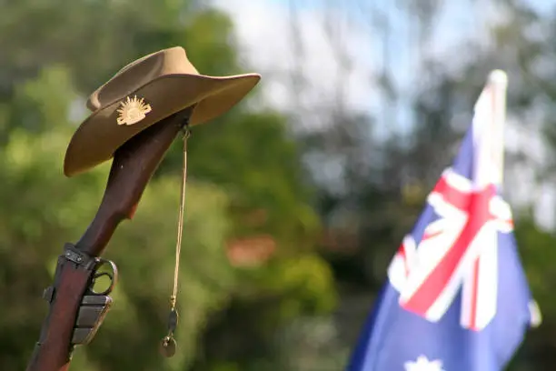 Landscape (with room for copy) of an upturned, vintage Australian Army 303 rifle (focus on the butt, trigger and magazine), a soldier's dogtags, iconic Aussie slouch hat (clearly showing the rising sun badge). The Australian National Flag can be seen flying in the background with sunlight passing through it. This image could be used to suit a range of creative or editorial contexts relating to Australian cultural identity, role in world military history or to simply honour those who have been injured or fallen in war, or have served in the military in peacetime, peacekeeping missions or during wartime.