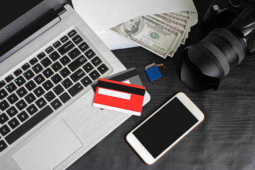 Top veiw of laptop, smartphone, camera, credit card, pen, notebook, dollars on the black wooden table