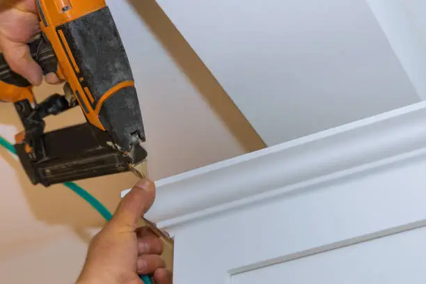 Air nailer tool close up of carpenter using nail gun to crown moldings on kitchen cabinets with white cabinets