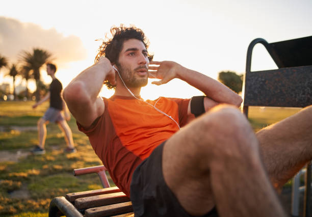 Confident young man listening to music in headphones from smart phone armband doing exercises on public equipment in the outdoor gym at the park Fitness young man doing exercises on public equipment in the park sit ups stock pictures, royalty-free photos & images