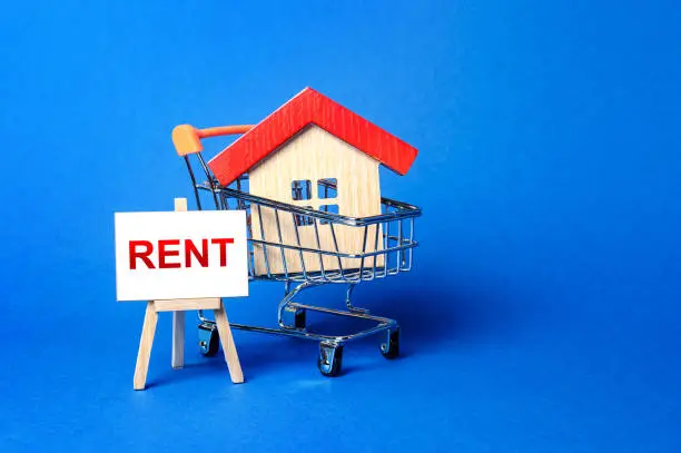 Photo of A house in a shopping cart and an easel with a word Rent. Rental housing and apartments, choice between buying and renting. Real estate prices and the desirability of buy a home or apartment.