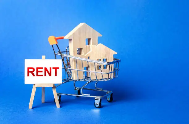 Photo of Houses in a shopping cart and an easel with a word Rent. Rental housing and apartments, choice between buying and renting. Real estate prices and the desirability of buy a home or apartment.