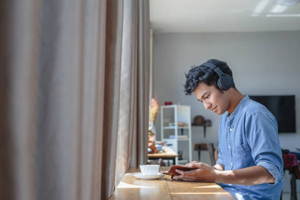 joven asiático guapo hombre escuchando música con auriculares y leer un libro en casa, personas relajante estilo de vida y concepto de tecnología - asian cuisine audio fotografías e imágenes de stock