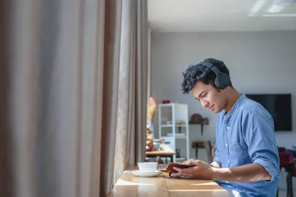 Photo of Young Asian handsome man listening to music with headphone and reading a book at home, People relaxing lifestyle and technology concept