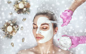 A doctor does face mask the skin to beautiful woman. Next to her are Christmas decorations. There are snowflakes in the background. New Year's and Cosmetology concept.