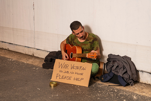 Photo of Homeless War Veteran who is Sitting in the Train Station, Tunnel or Underground and Playing Guitar with Help Sign and Box for Donation. Desperate Mature Man is Feeling Abandoned and Lost in Depression and Sitting on Ground Street While Suffering Emotional Pain.