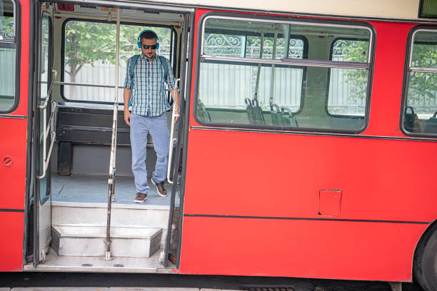 Handsome Man in Plaid Shirt and with Backpack is Exiting the Public Bus and Listening to Music. Casually Dressed Tourist with Sunglasses and Headphones at the Doors of a Public Transport.