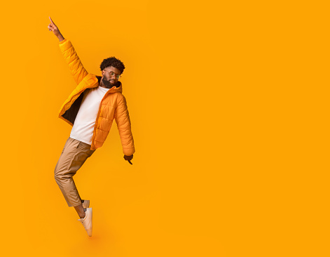 Joyful african man in winter clothes posing over orange background, standing on toes with one hand up, free space