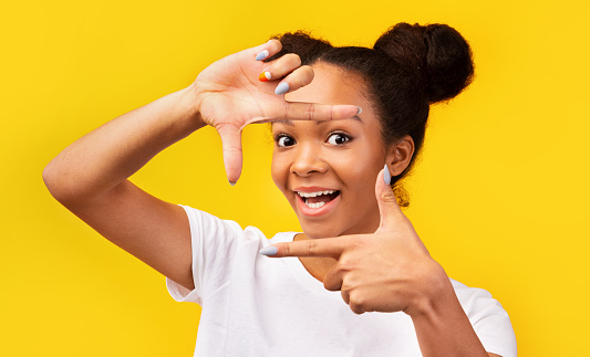 Close-up Portrait Of Cheerful Black Girl Making Frame Using Her Hands And Fingers, Isolated On Yellow Background